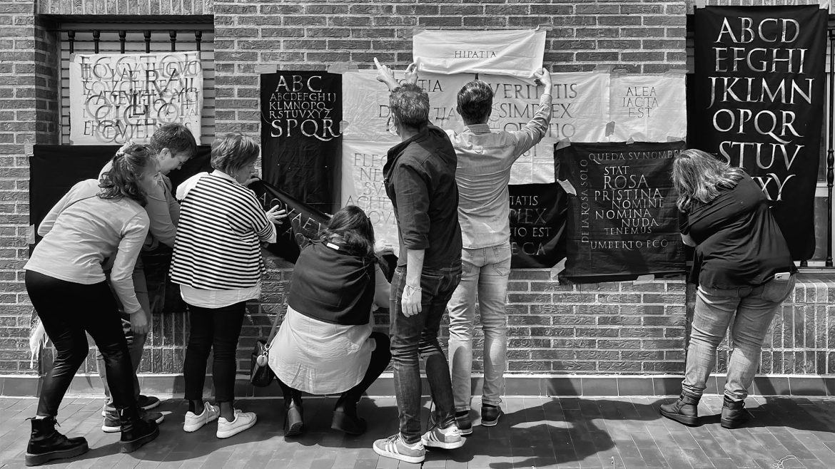 Grupo de alumnos del curso de caligrafía colocando sus trabajos finales de alfabetos romanos y capitulares en una pared de ladrillo durante una exposición.