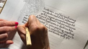 Recreación de Manuscrito con Letra Capital Iluminada en escritura Carolingia Primer plano de unas manos escribiendo un texto en latín con plumilla plana junto al boceto a lápiz de una gran letra capital decorada estilo románico en un taller de Asturias Caligrafía.