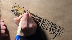 Práctica de Alfabeto con Pluma Brause Primer plano de una mano practicando el alfabeto con tinta amarilla sobre letras negras utilizando una plumilla Brause en un taller de Asturias Caligrafía.