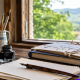 Mesa de madera de Asturias Caligrafía con pluma estilográfica, tintero, pinceles y láminas de caligrafía junto a una ventana con vistas al paisaje de Asturias.